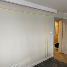 PM Remonty i Konserwacja Obiektów - Przygotowanie do malowania: ściana z taśmą malarską, poziomica laserowa wyznacza linię. Podłoga zabezpieczona folią. Widoczne gniazdka elektryczne i fragment parkietu.