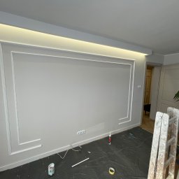 PM Remonty i Konserwacja Obiektów - Szara ściana z dekoracyjną białą ramą i oświetleniem LED. Podłoga zabezpieczona folią, widoczna drabina i akcesoria malarskie. Detal wykończenia wnętrza.