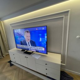 PM Remonty i Konserwacja Obiektów - Elegancki salon z telewizorem w ozdobnej ramie ściennej i podświetleniem LED. Jasne meble i parkiet tworzą nowoczesne wnętrze. Widoczne gniazdka elektryczne.
