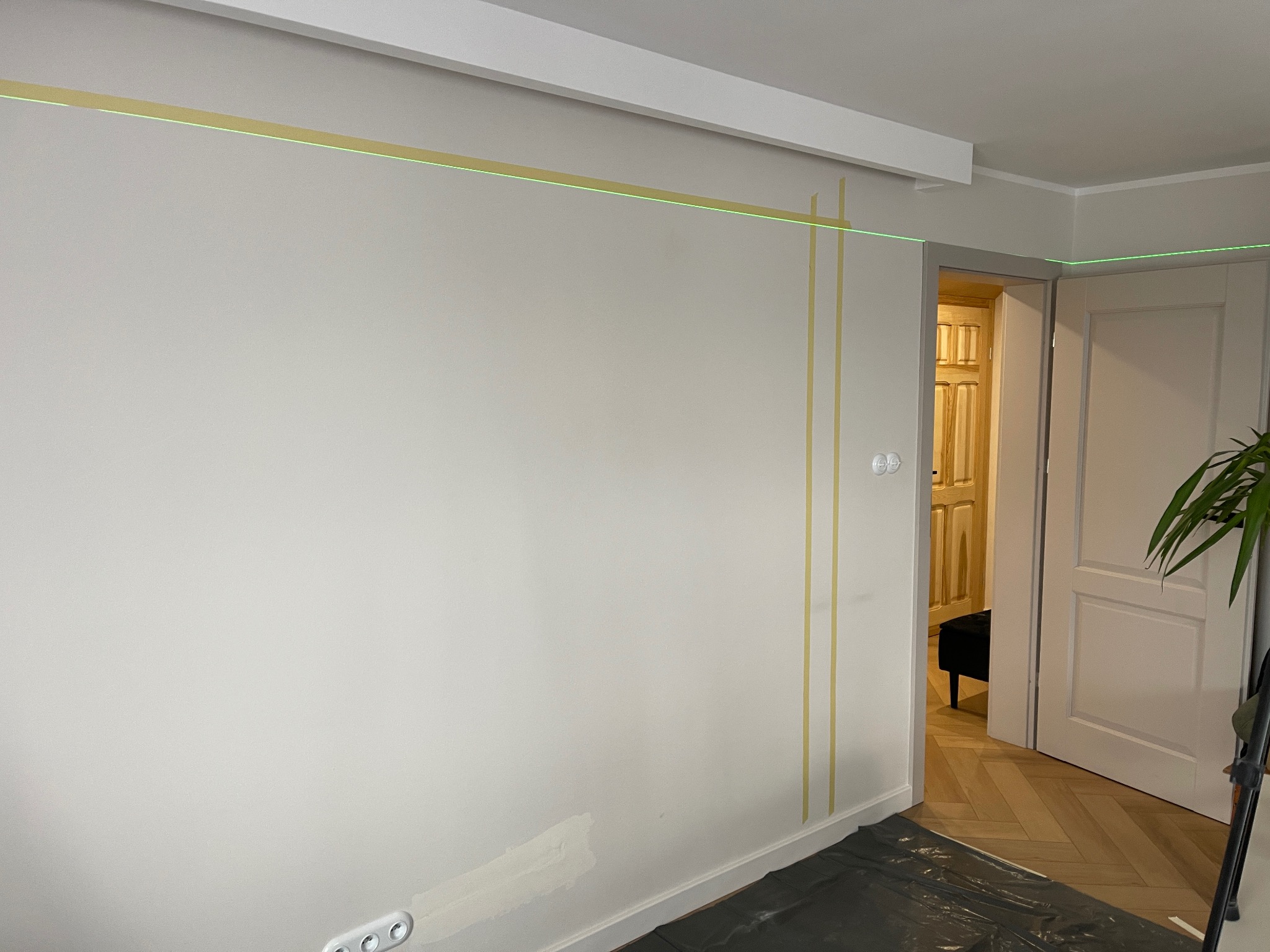 Przygotowanie do malowania: ściana z taśmą malarską, poziomica laserowa wyznacza linię. Podłoga zabezpieczona folią. Widoczne gniazdka elektryczne i fragment parkietu.