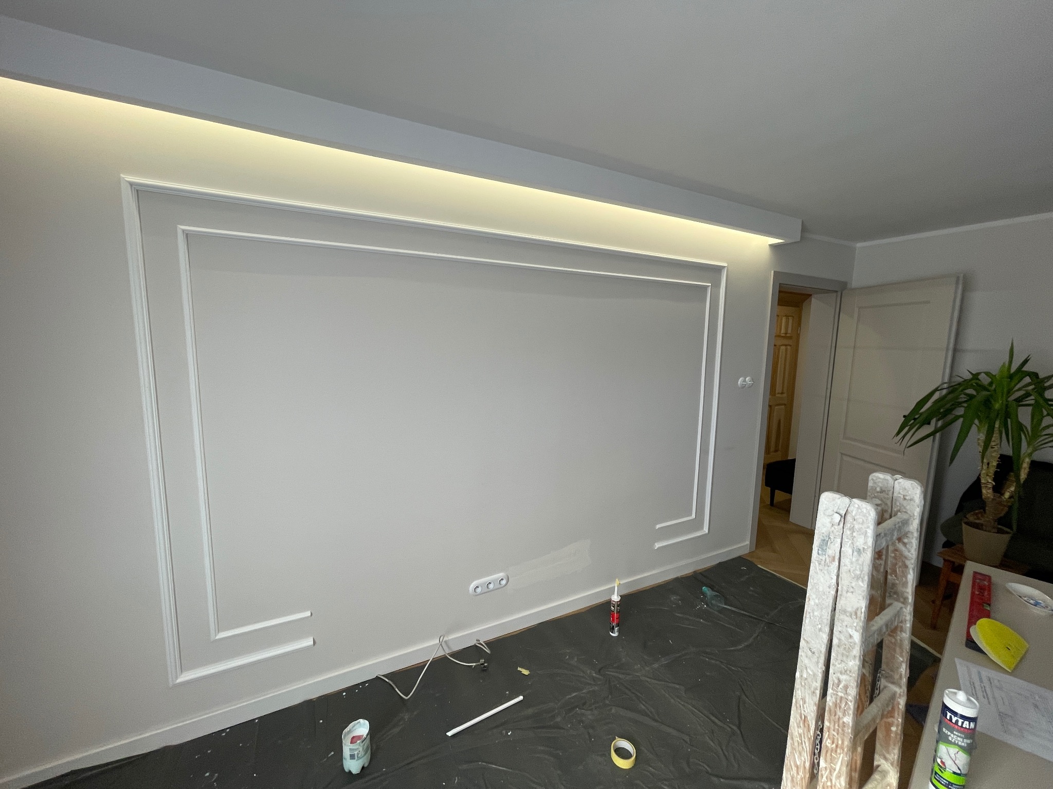 Szara ściana z dekoracyjną białą ramą i oświetleniem LED. Podłoga zabezpieczona folią, widoczna drabina i akcesoria malarskie. Detal wykończenia wnętrza.