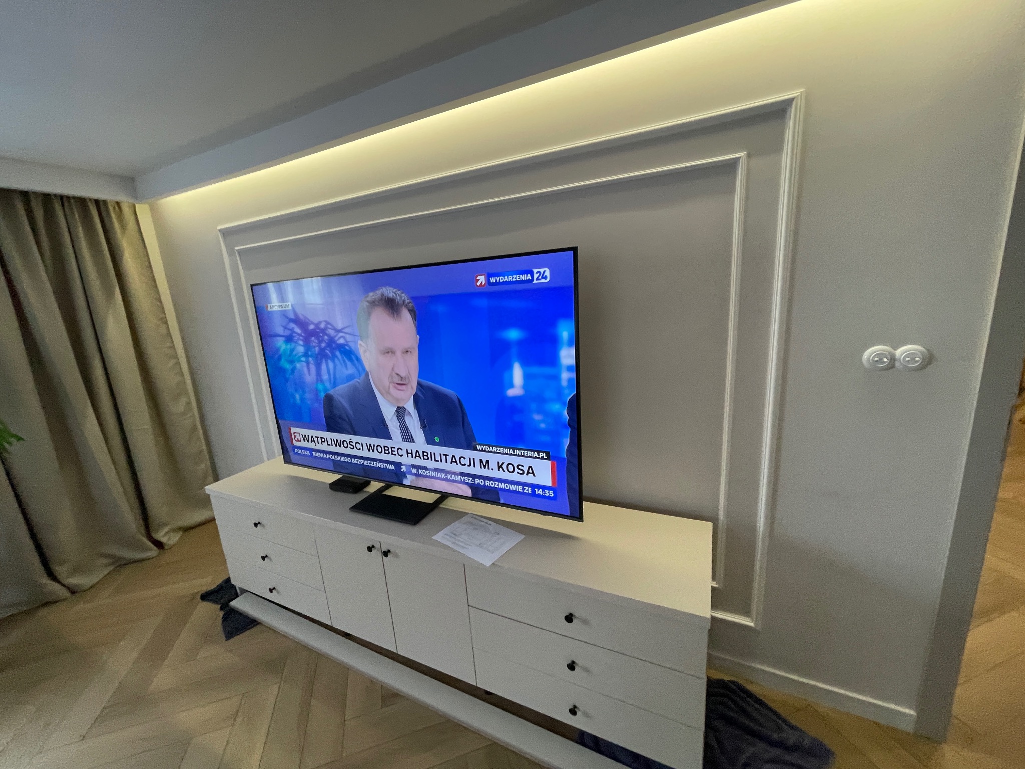Elegancki salon z telewizorem w ozdobnej ramie ściennej i podświetleniem LED. Jasne meble i parkiet tworzą nowoczesne wnętrze. Widoczne gniazdka elektryczne.