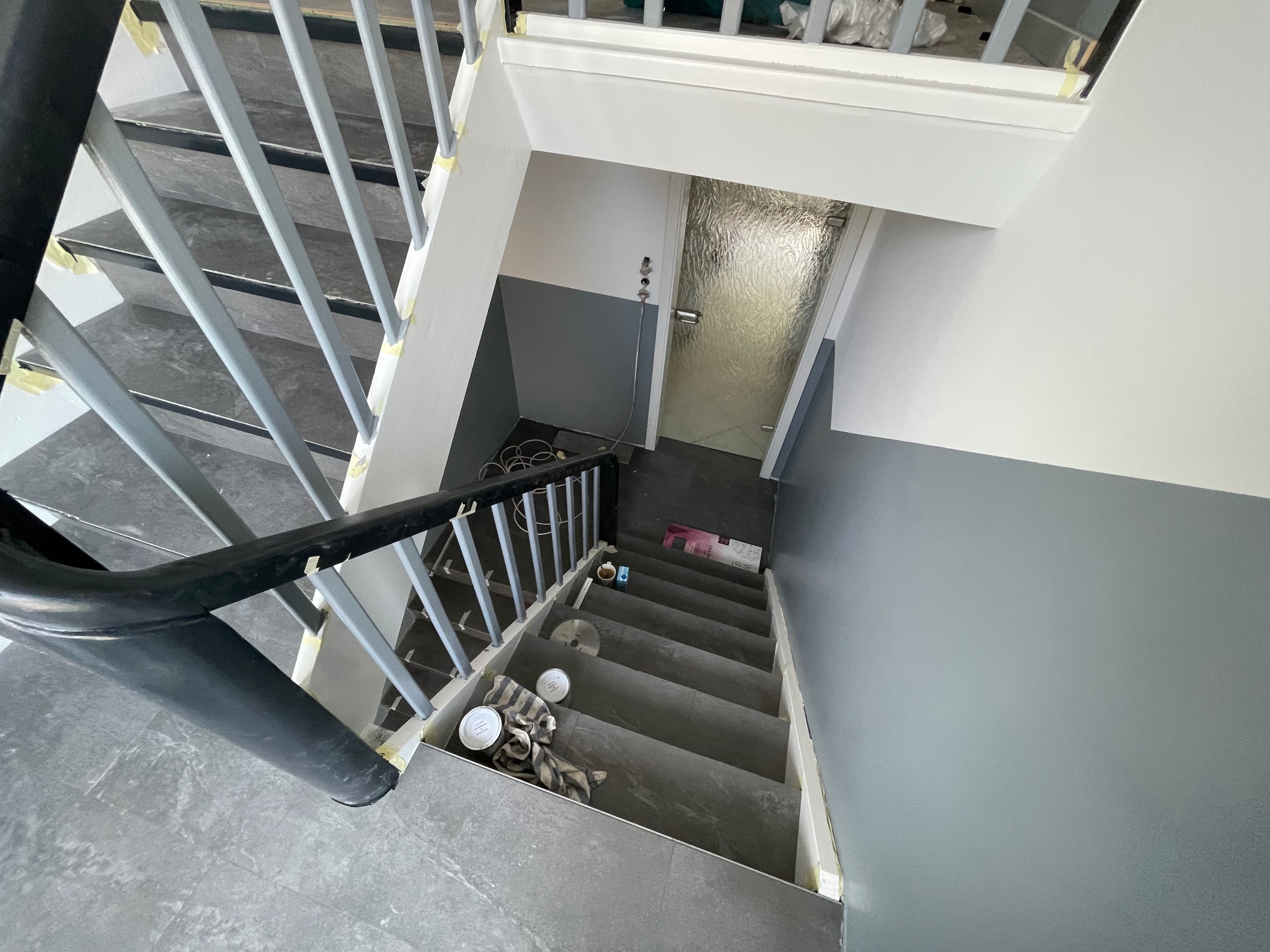 Klatka schodowa w trakcie malowania: szare ściany do połowy wysokości, białe powyżej. Zabezpieczone taśmą schody i poręcze. Widoczne puszki z farbą i narzędzia.