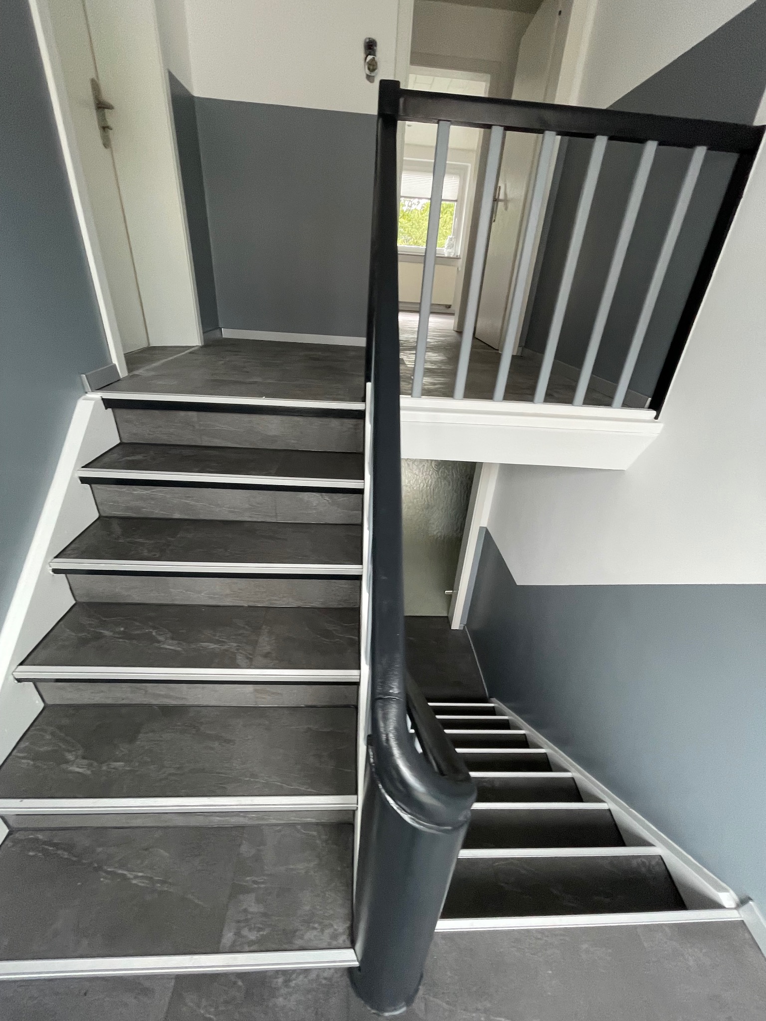 Wykończone schody z szarymi płytkami imitującymi kamień, białe stopnice i szara ściana z białym pasem u góry. Nowoczesna balustrada z szarymi prętami.