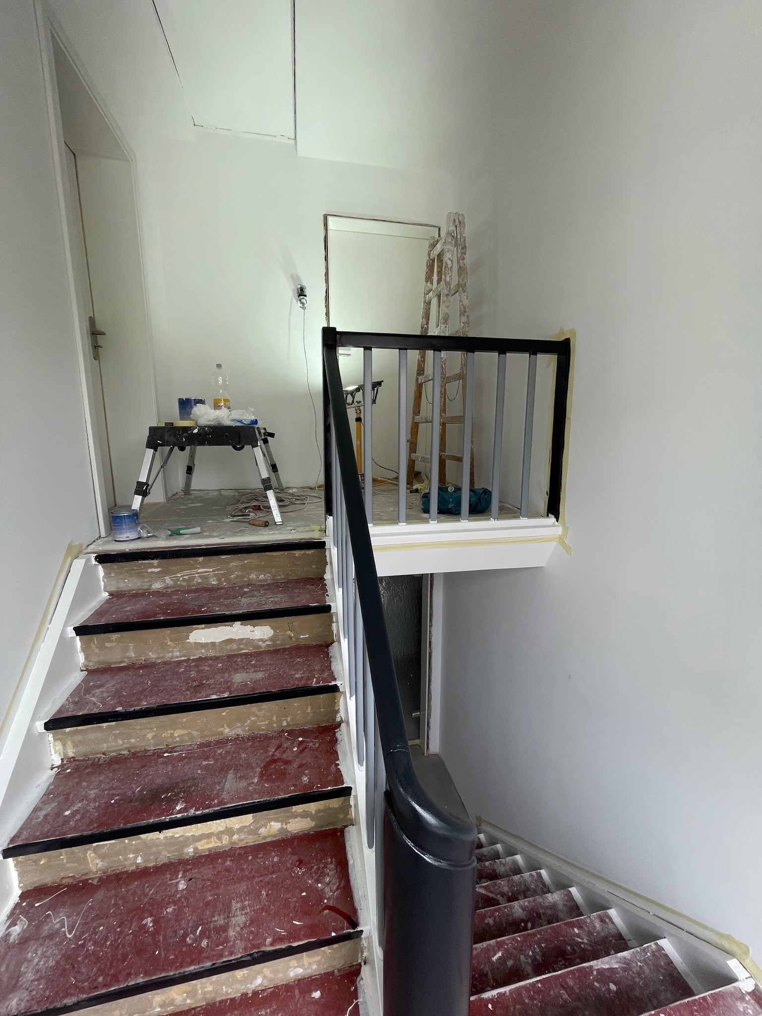 Klatka schodowa w trakcie malowania: zabezpieczone taśmą schody z resztkami farby, drabina i narzędzia malarskie w tle. Widok z góry na dół.