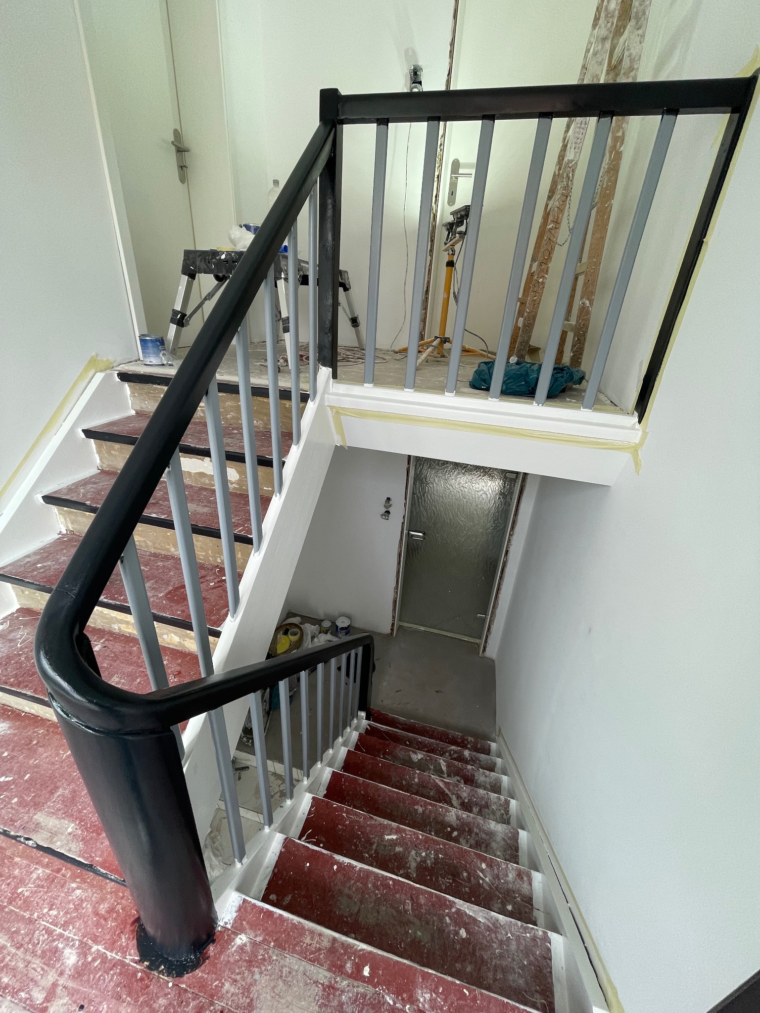 Malowanie klatki schodowej w trakcie remontu. Widoczne zabezpieczenia taśmą malarską, pomalowane barierki i schody z resztkami farby. W tle drabina i narzędzia.