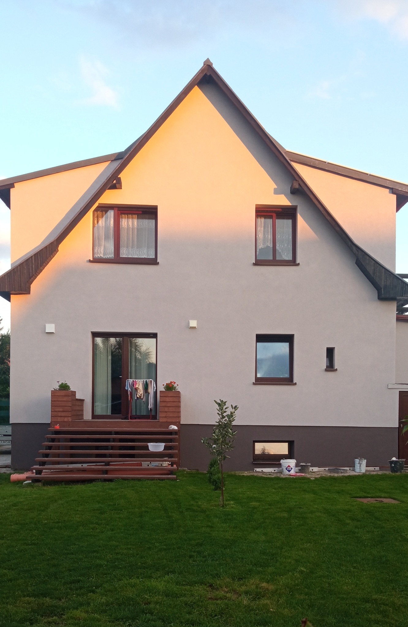 Dwukondygnacyjny dom z jasną elewacją i brązowymi akcentami, widoczny w popołudniowym świetle, z elementami wskazującymi na niedawne prace malarskie: wiaderka z farbą i drabina leżąca na trawie...