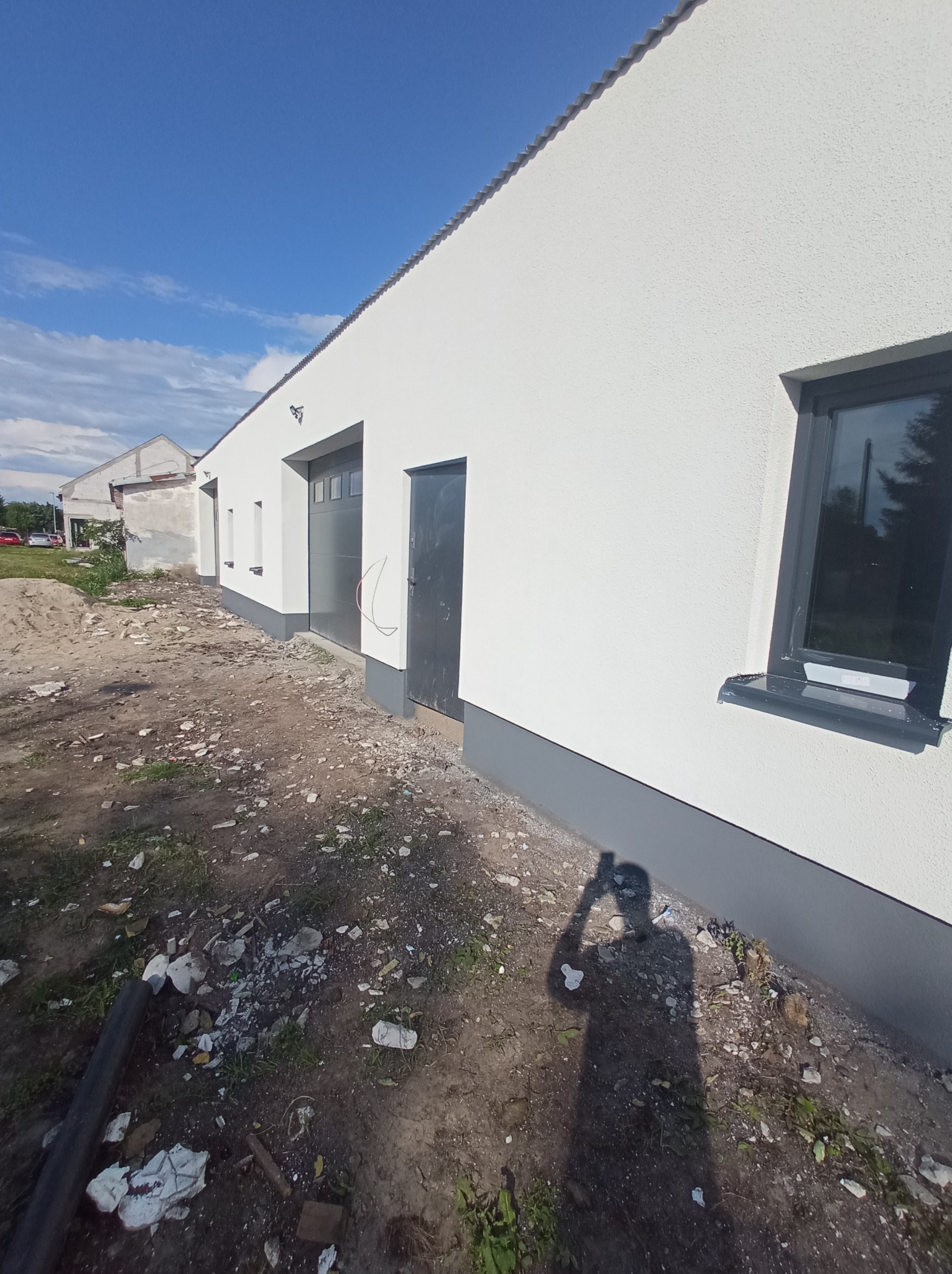 Świeżo otynkowana, biała elewacja budynku gospodarczego z szarymi elementami wykończeniowymi, widoczny fragment niezagospodarowanego terenu z gruzem.