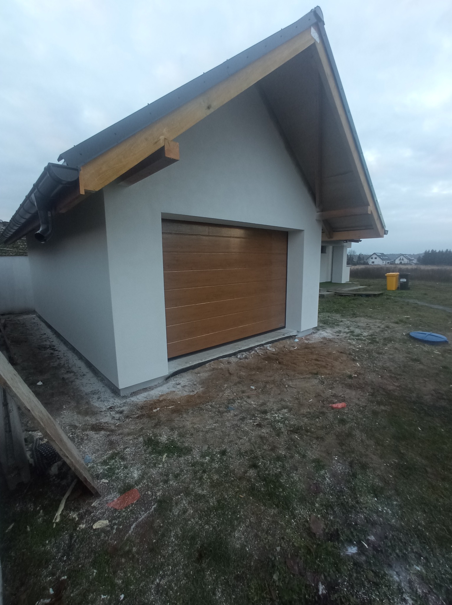 Nowo otynkowany garaż z brązową bramą segmentową, widoczne ślady prac budowlanych na trawie, pochmurne niebo.