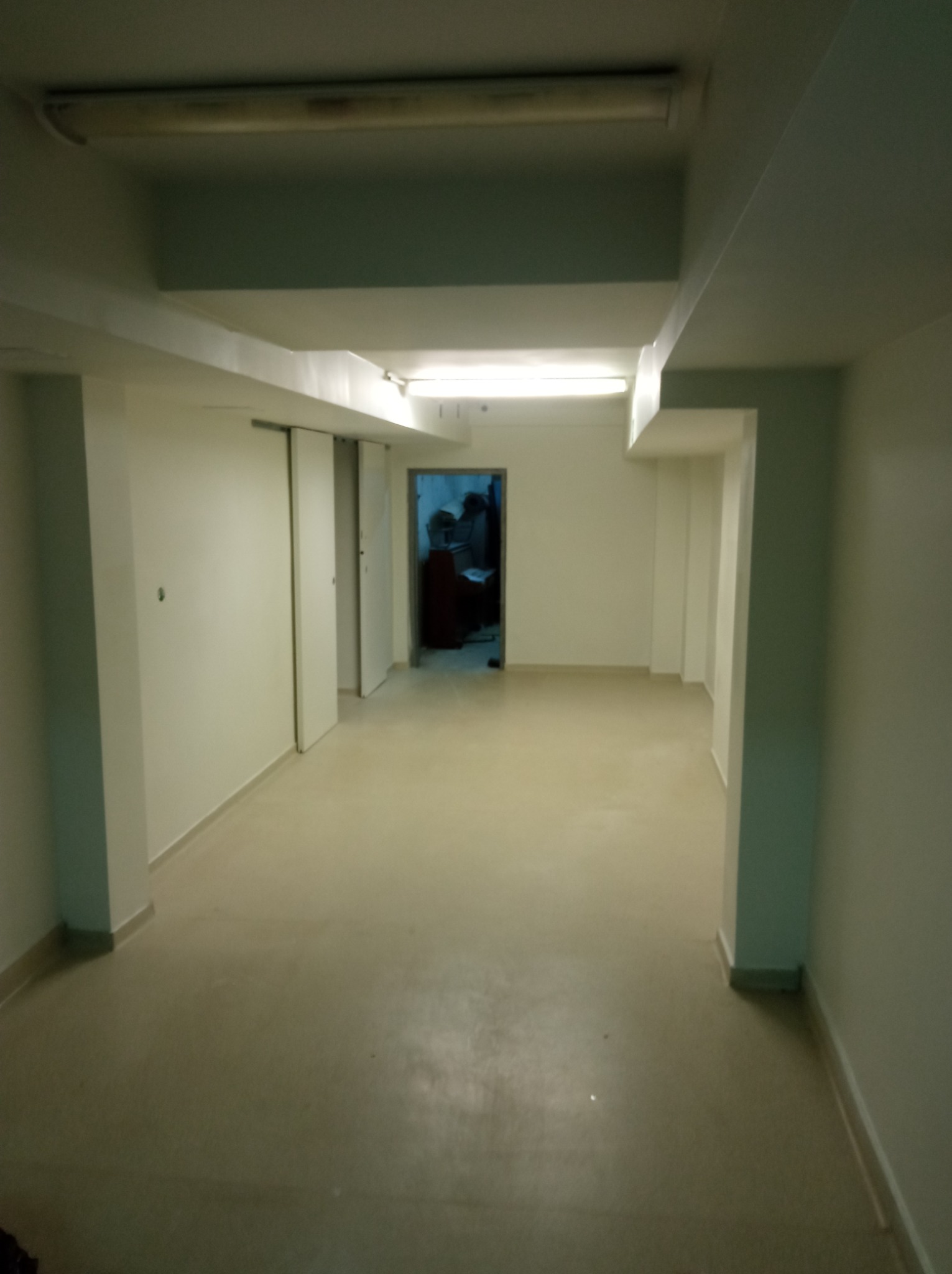 Pusta, świeżo pomalowana korytarz w piwnicy z białymi ścianami, szarą podłogą i widocznymi elementami konstrukcyjnymi pod sufitem. W głębi korytarza otwarte drzwi prowadzą do pomieszczenia...