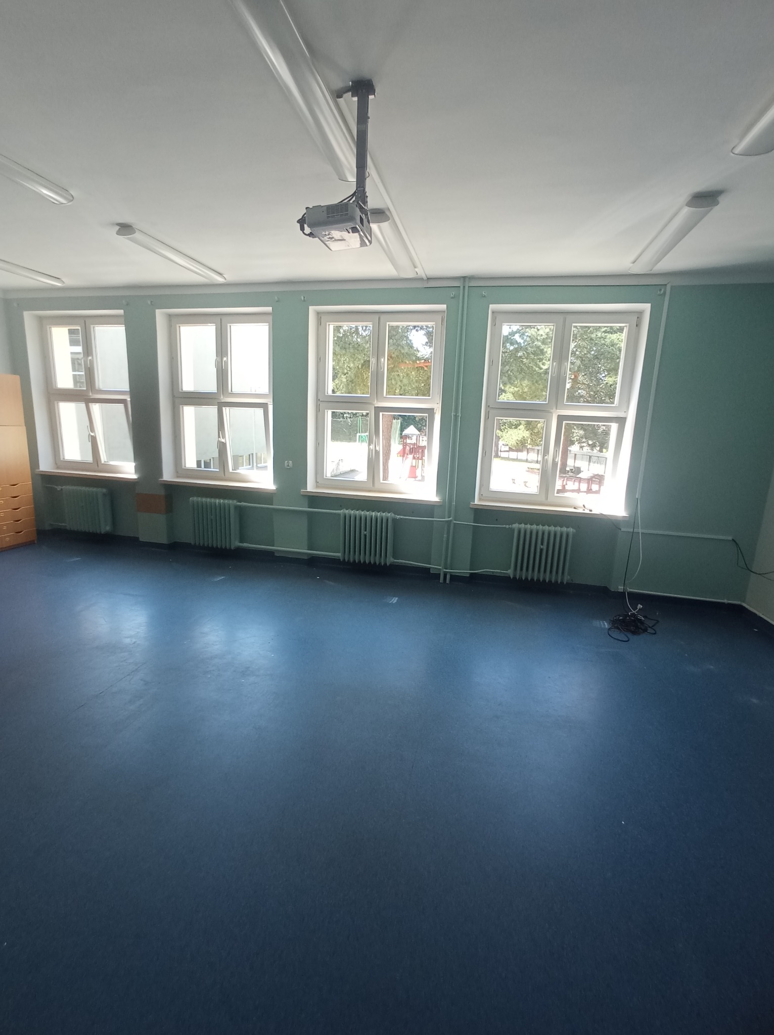 Puste pomieszczenie z niebieską podłogą, trzema oknami z widokiem na plac zabaw, zielonymi ścianami i grzejnikami pod oknami, oświetlone lampami sufitowymi i wiszącym projektorem.