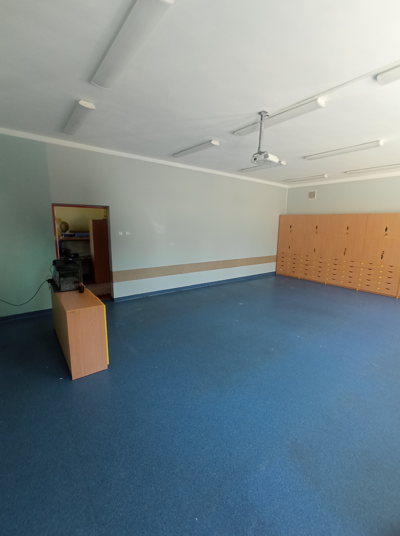 Wnętrze pustej sali szkolnej z niebieską podłogą, rzędem drewnianych szafek i projektorem na suficie.