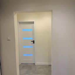 SERHII HULIAVSKYI sh.remontpro - Białe drzwi z poziomymi, błękitnymi przeszkleniami w jasnym wnętrzu. Szare płytki podłogowe i kremowe ściany. Widoczny fragment korytarza z włącznikiem światła.