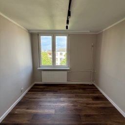 SERHII HULIAVSKYI sh.remontpro - Wnętrze pokoju po remoncie: ciemne panele podłogowe, świeżo pomalowane ściany w odcieniach beżu, białe listwy przypodłogowe i okno z widokiem na zieleń.