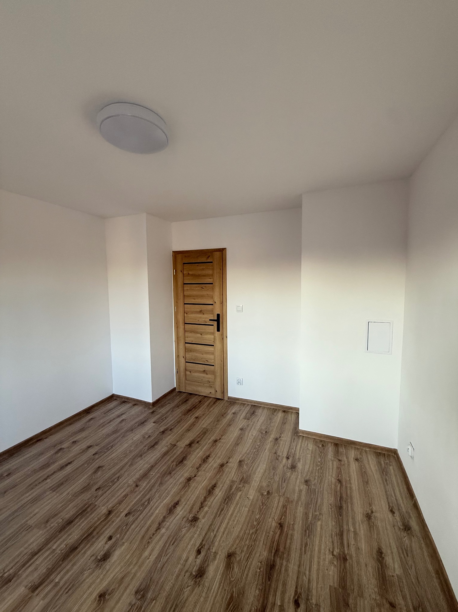 Jasne wnętrze po remoncie: drewnopodobne panele, białe ściany z gładzią, drewniane drzwi z czarną klamką i okrągła lampa sufitowa. Widok na wykończony pokój.