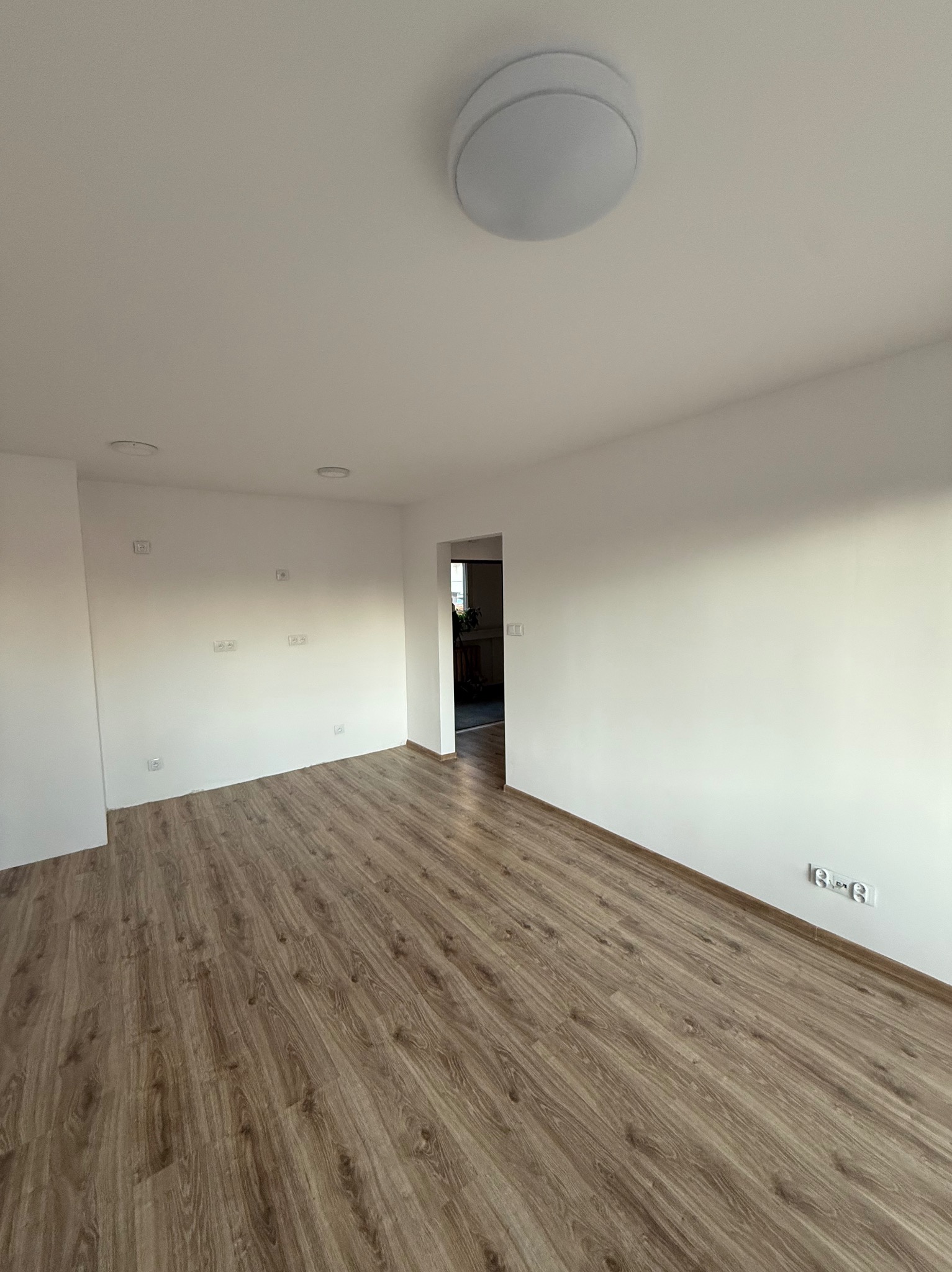 Jasne wnętrze po remoncie: panele podłogowe w odcieniach brązu, białe ściany z gniazdkami elektrycznymi, sufit z okrągłą lampą. Widok na sąsiednie pomieszczenie.