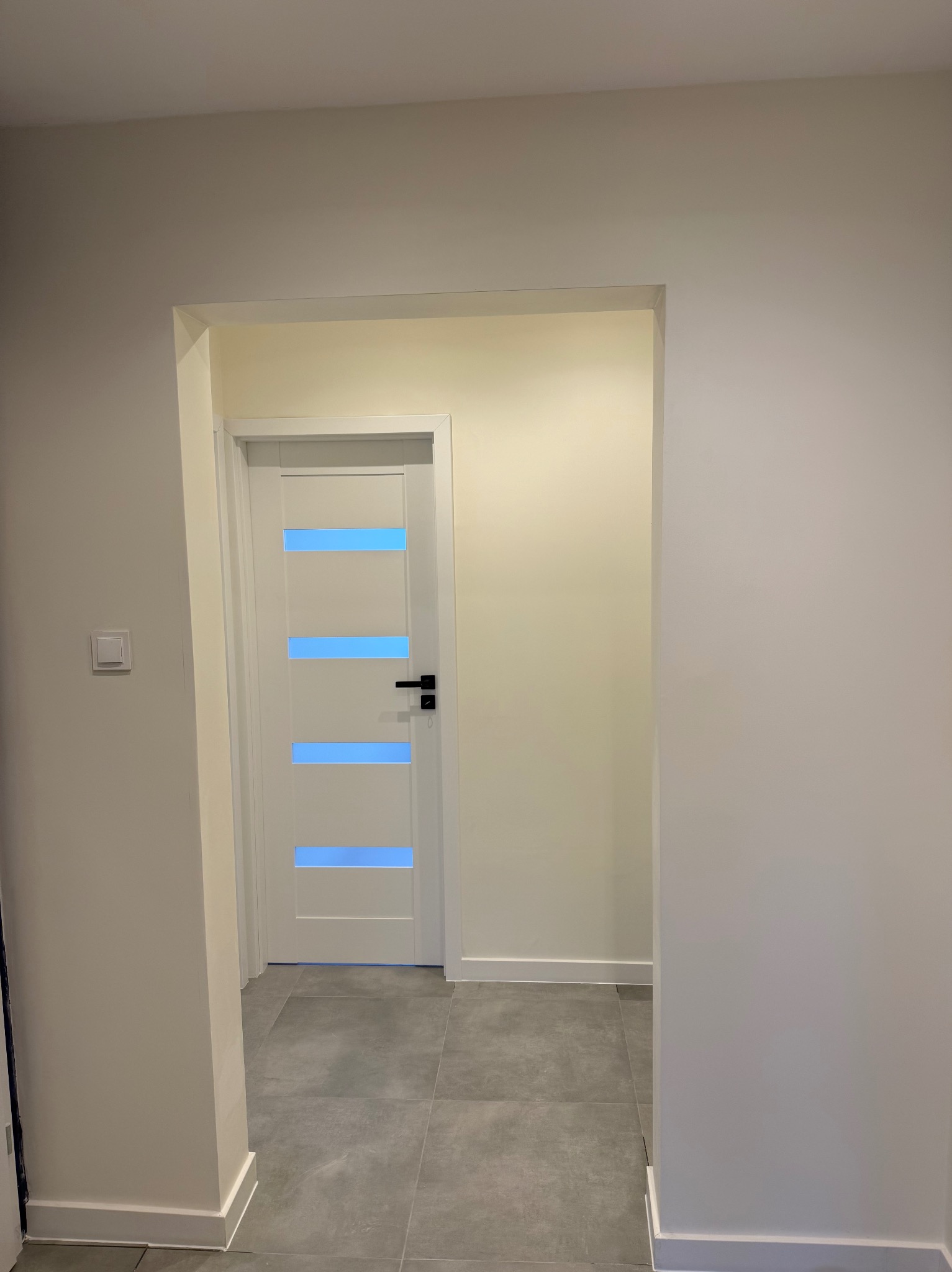 Białe drzwi z poziomymi, błękitnymi przeszkleniami w jasnym wnętrzu. Szare płytki podłogowe i kremowe ściany. Widoczny fragment korytarza z włącznikiem światła.