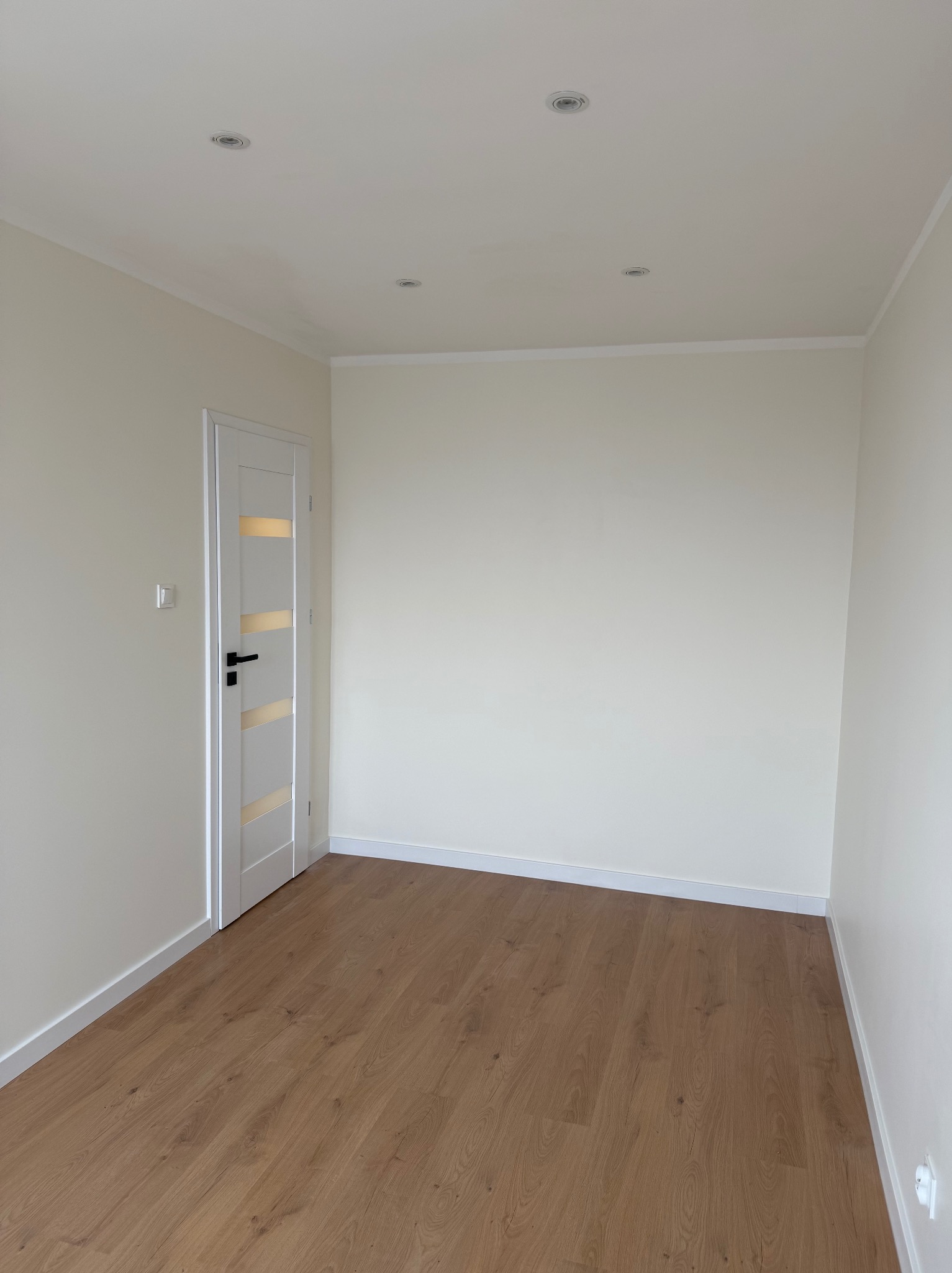 Jasne wnętrze po remoncie: drewniana podłoga, białe ściany z listwami, nowoczesne drzwi z przeszkleniami i punktowe oświetlenie sufitowe. Minimalistyczny design.