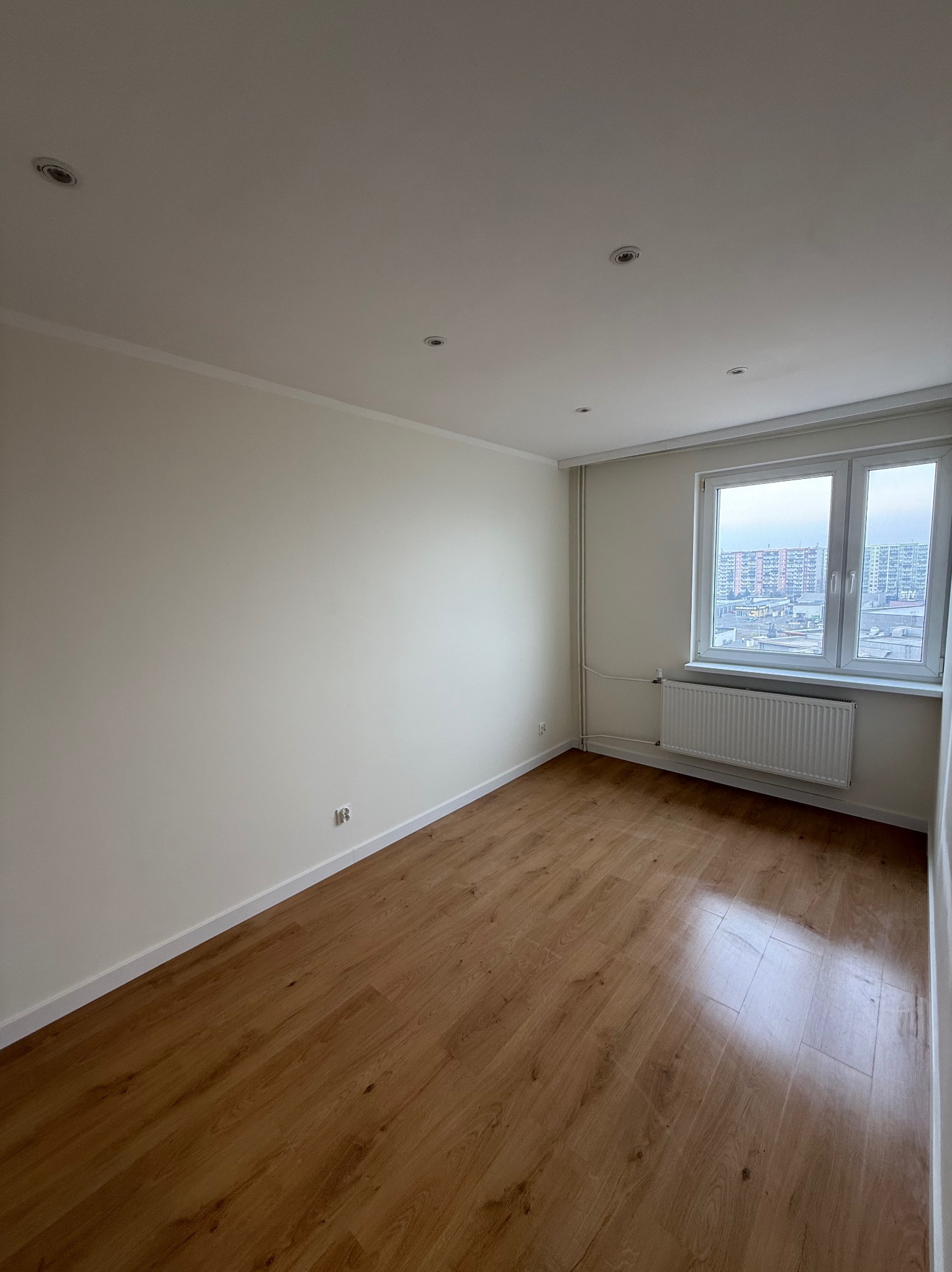 Jasne, puste pomieszczenie po remoncie: świeżo pomalowane ściany, ułożone panele podłogowe, okno z widokiem na miasto. Minimalistyczne wykończenie.