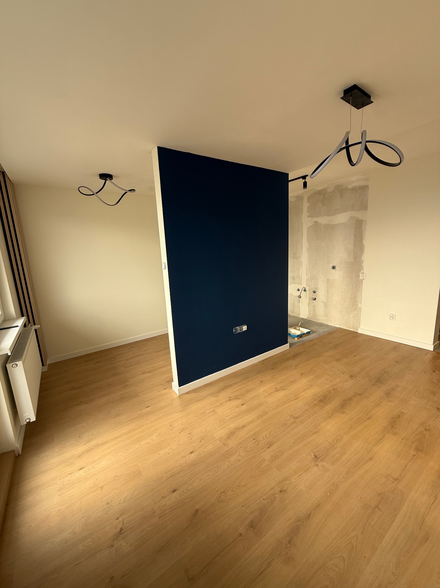 Pokój po remoncie: panele podłogowe, ściana pomalowana na granatowo, fragment ściany w szpachli, nowoczesna lampa. Widok na odnowione wnętrze z oknem.