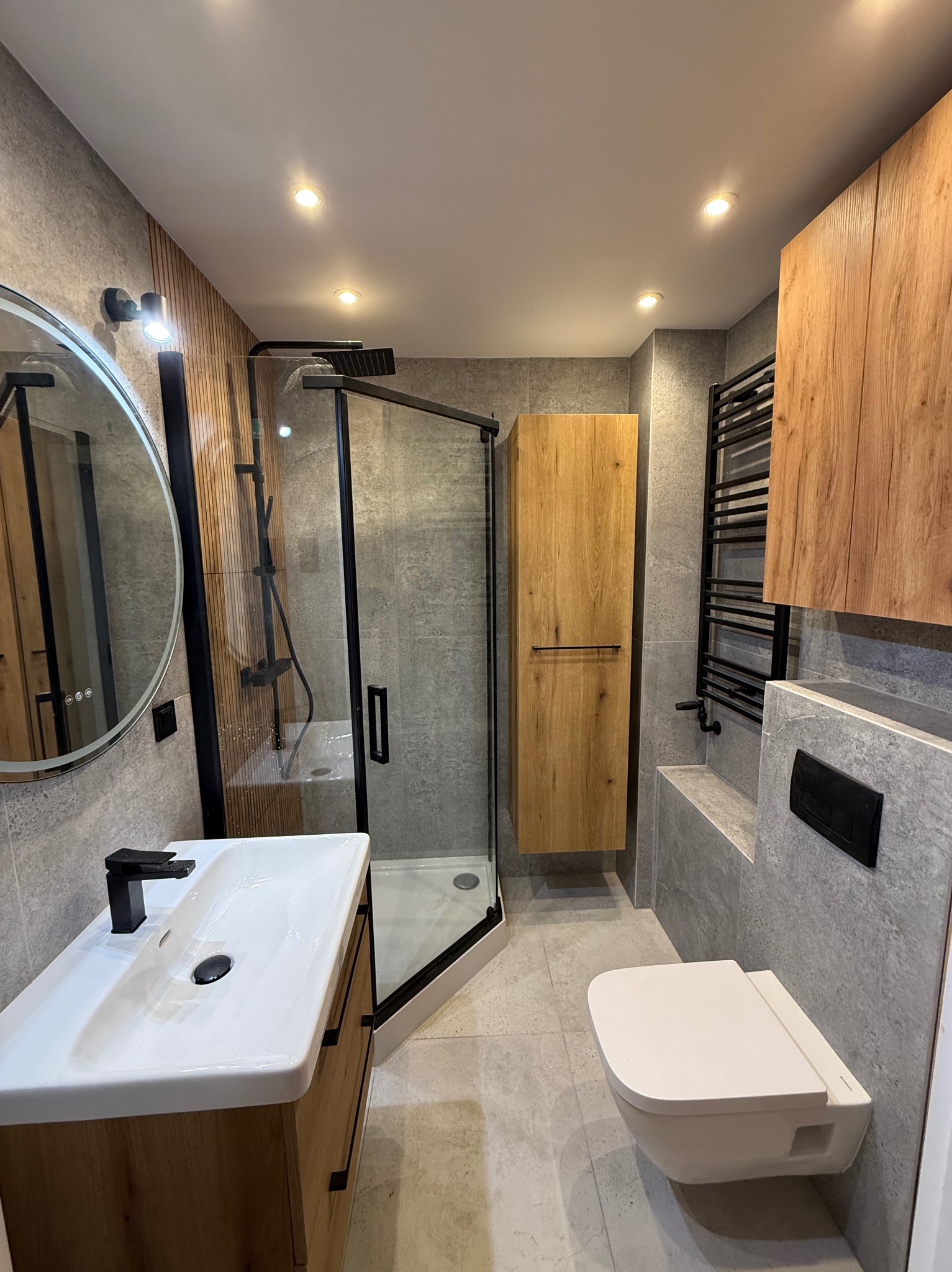 Nowoczesna łazienka z kabiną prysznicową typu walk-in, umywalką nablatową i drewnianymi szafkami. Szare płytki imitujące kamień na ścianach i podłodze. Czarne akcenty.