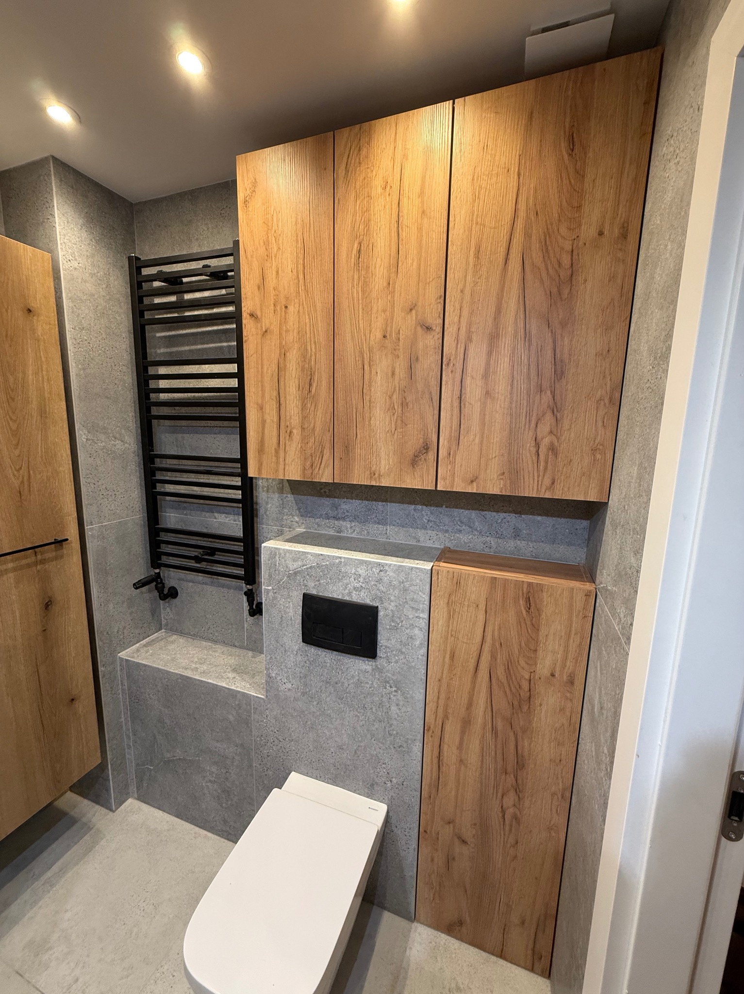 Nowoczesna łazienka po remoncie: szare płytki, drewniane szafki, czarny grzejnik drabinkowy i biała toaleta. Minimalistyczny design z dbałością o detale.