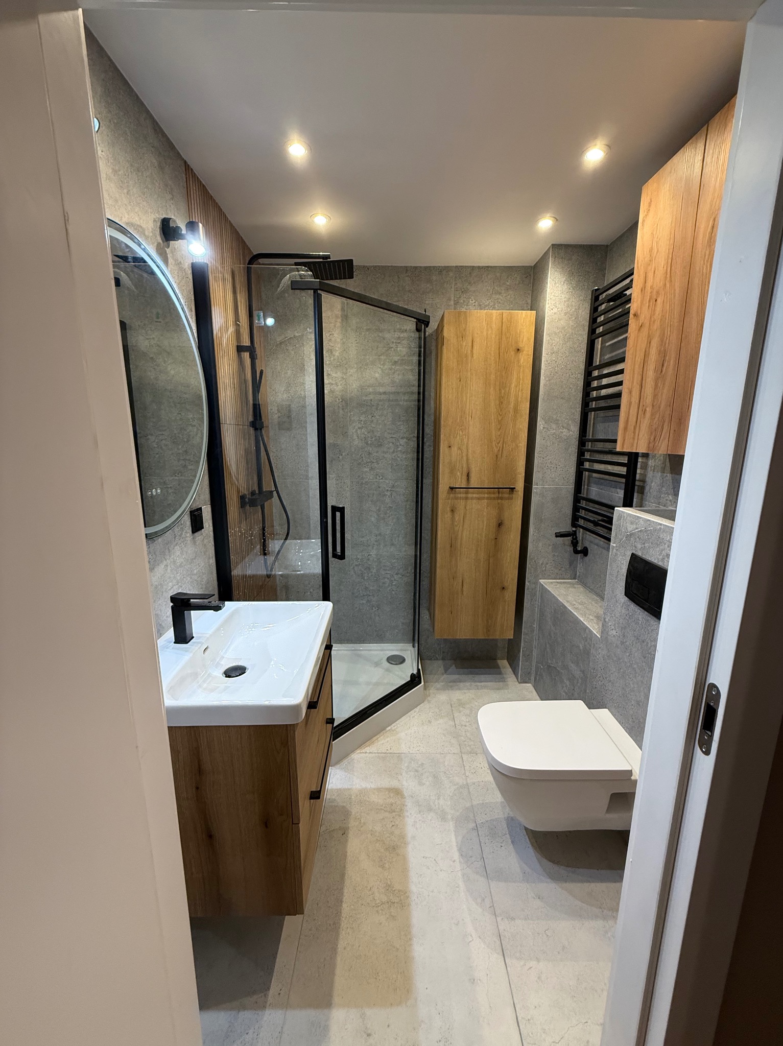 Nowoczesna łazienka z kabiną prysznicową typu walk-in, umywalką z szafką z drewnianym fornirem i wiszącą toaletą. Szare płytki i czarne akcenty.