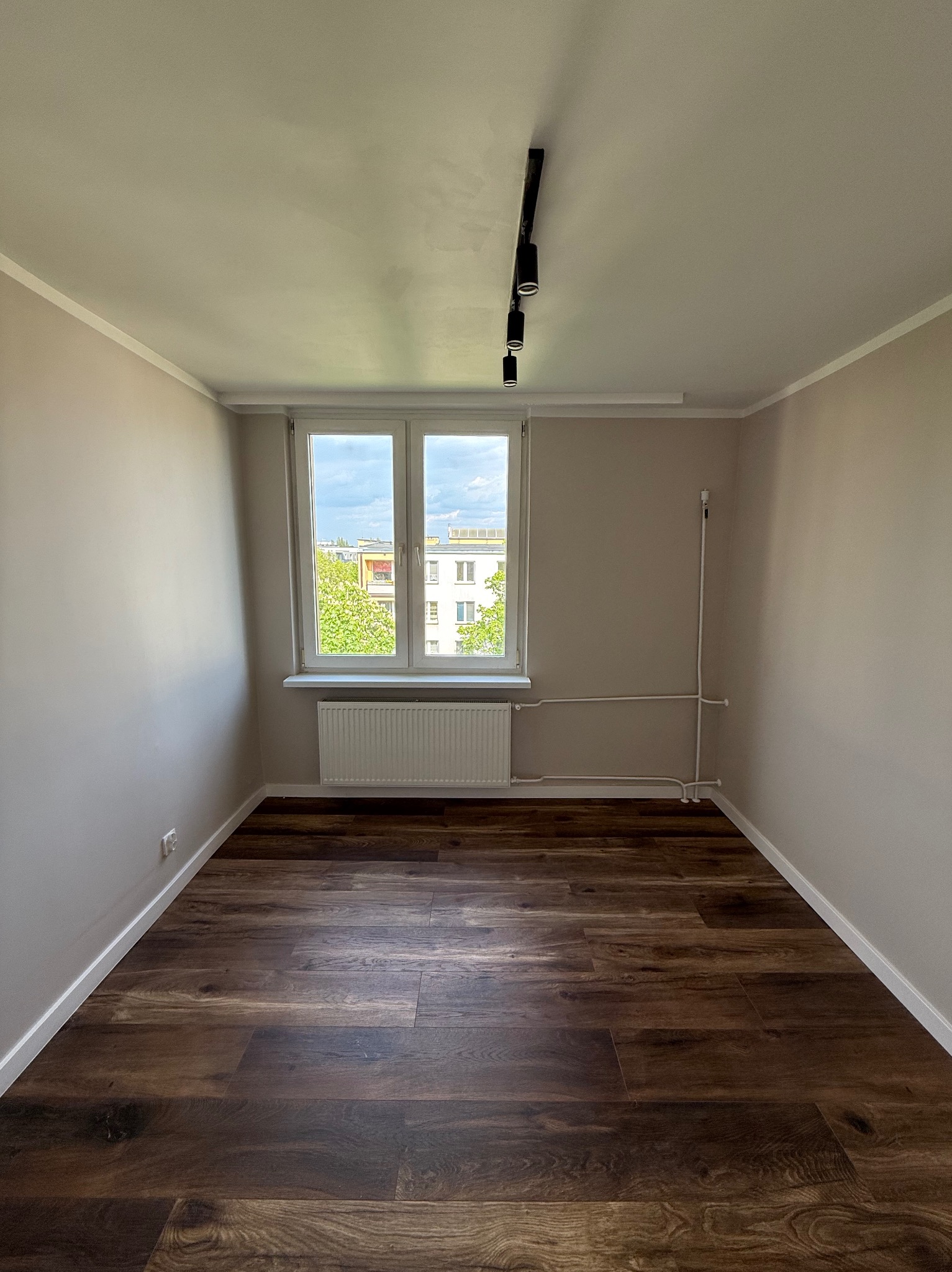 Wnętrze pokoju po remoncie: ciemne panele podłogowe, świeżo pomalowane ściany w odcieniach beżu, białe listwy przypodłogowe i okno z widokiem na zieleń.