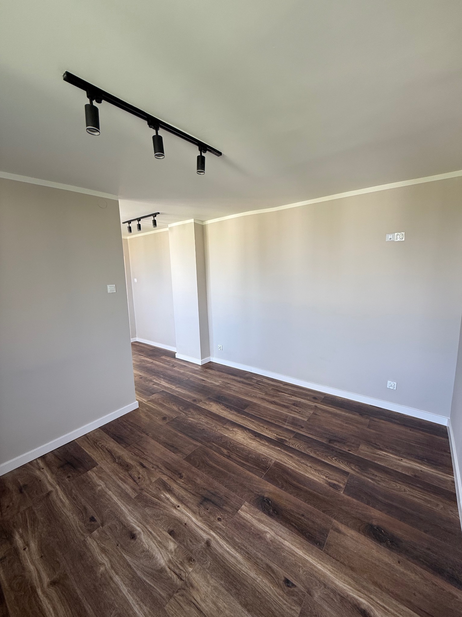 Wykończone wnętrze: brązowe panele podłogowe, beżowe ściany z białymi listwami, czarne oświetlenie szynowe. Widok na sąsiednie pomieszczenie.