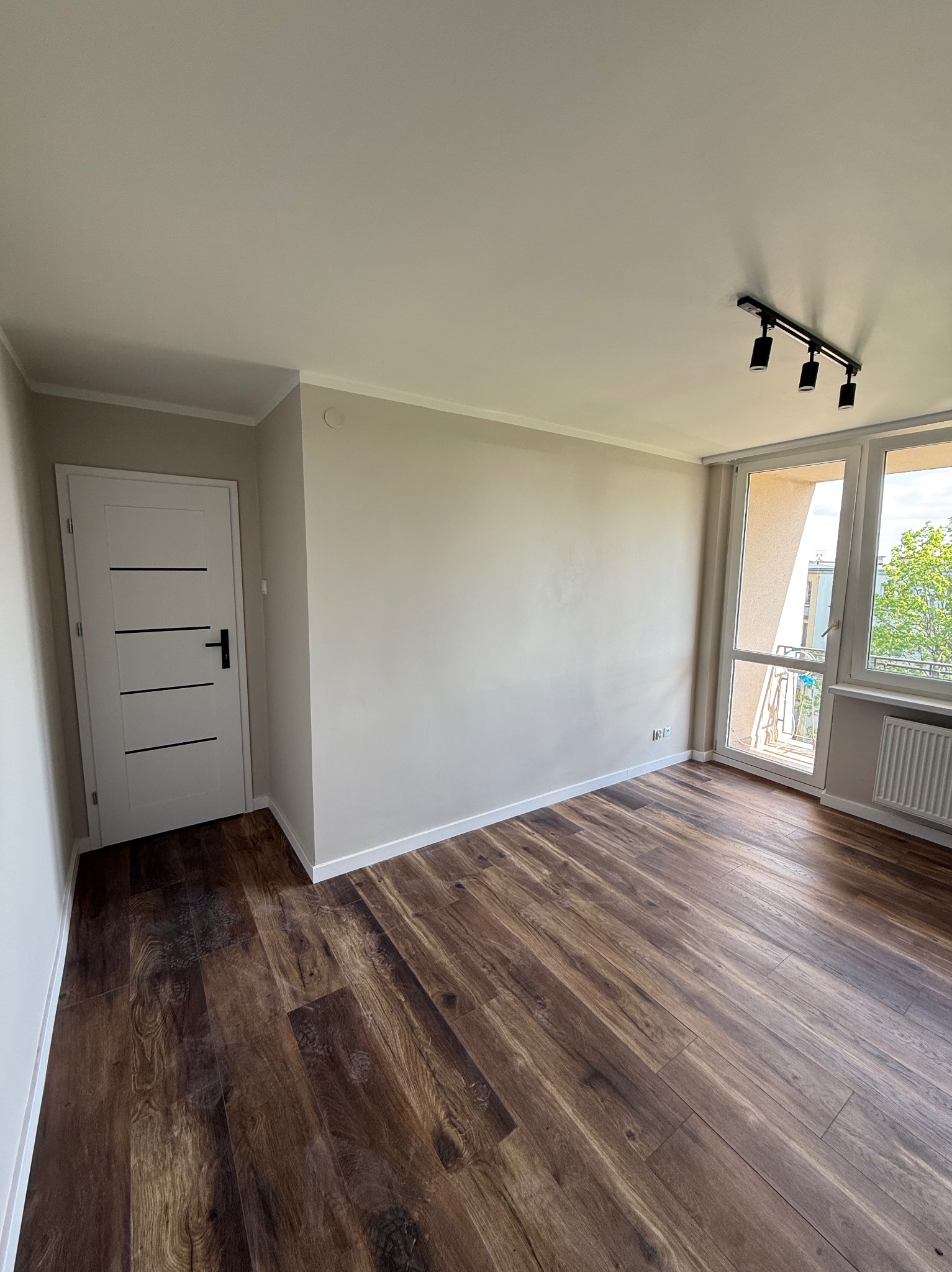 Jasne, odświeżone wnętrze po remoncie: ciemne panele podłogowe, kremowe ściany, białe drzwi z czarnymi akcentami i okno balkonowe z widokiem na zieleń.