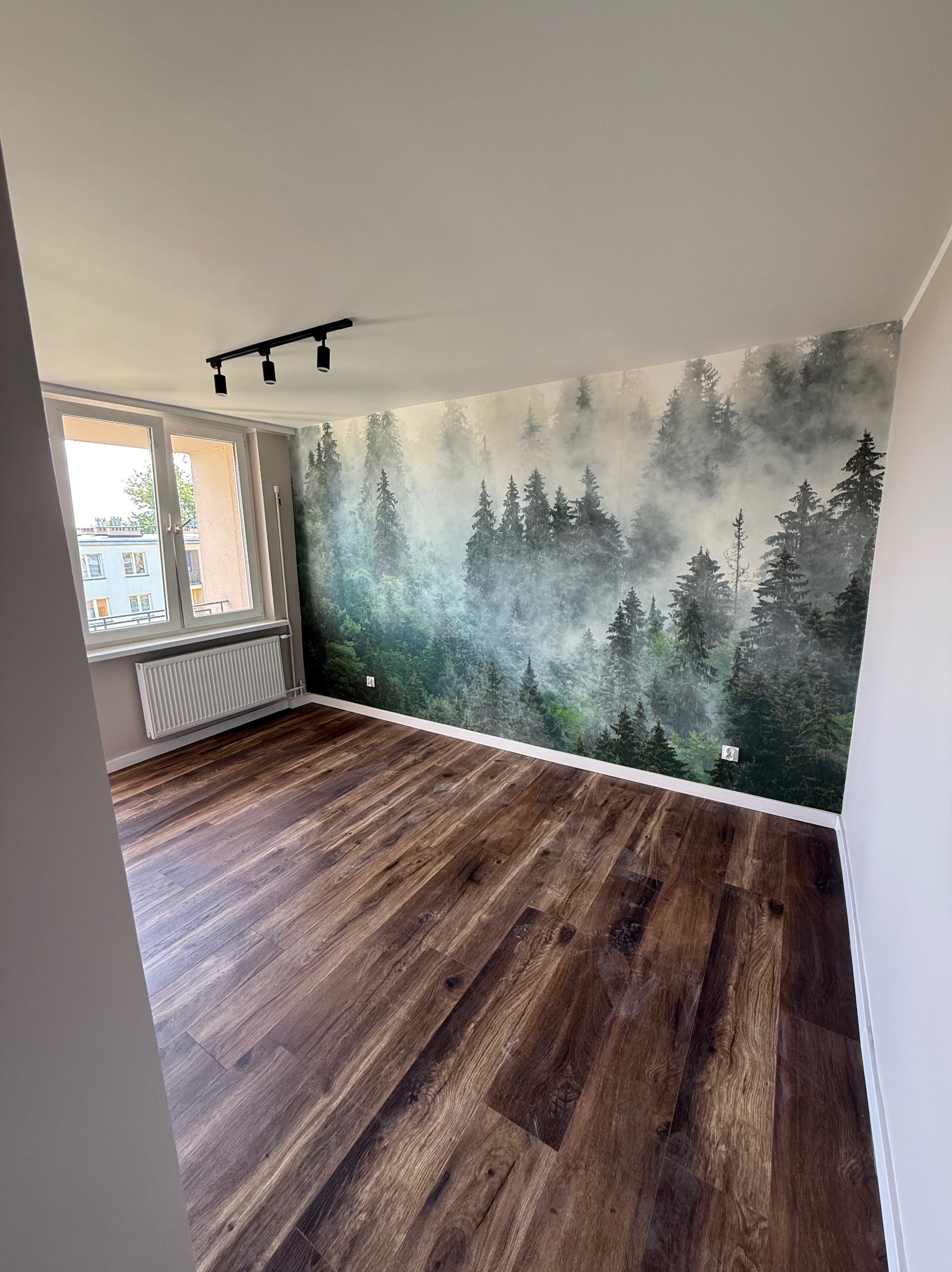 Pokój po remoncie: brązowe panele, ściana z fototapetą leśną, białe ściany i okno z grzejnikiem. Nowoczesne oświetlenie sufitowe.