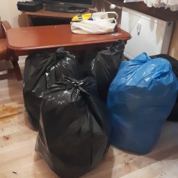 Mariusz Dziarnowski - Wnętrze mieszkania z czterema workami na śmieci, trzema czarnymi i jednym niebieskim, stojącymi na zabrudzonej podłodze. W tle widoczny telewizor na komodzie i grzejnik pod oknem.