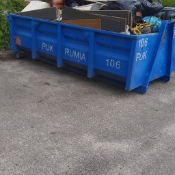 Mariusz Dziarnowski - Niebieski kontener PUK Rumia wypełniony odpadami: meblami, workami ze śmieciami i elementami wyposażenia wnętrz, na tle drzew i chodnika.