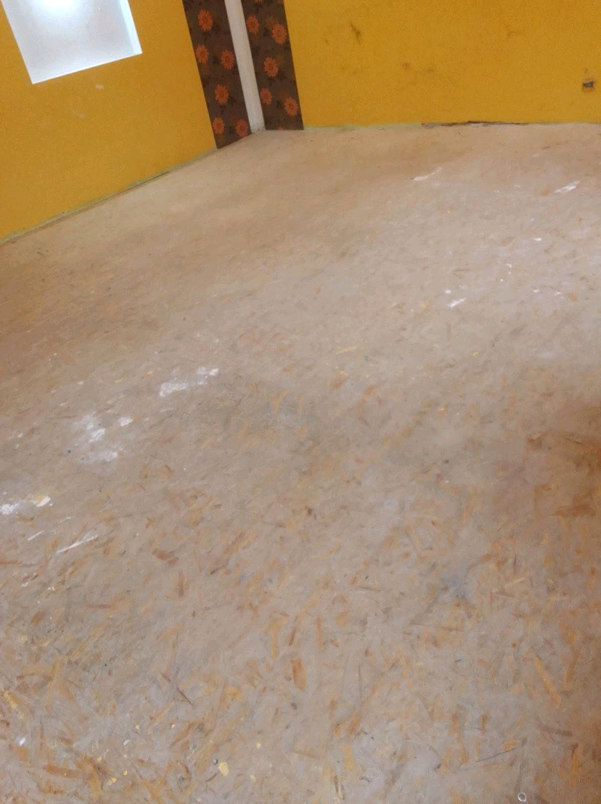 Pusty pokój z żółtymi ścianami i podłogą z płyty OSB, ślady po remoncie na podłodze, widok z góry, dekoracyjny pas tapety na rogu ściany.