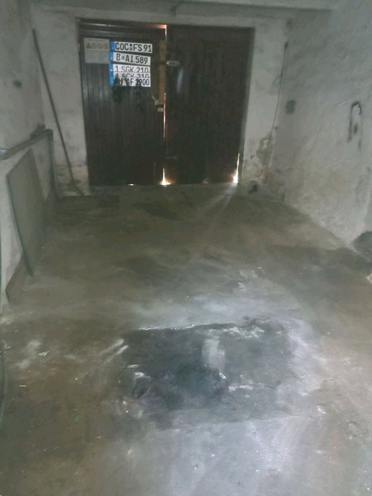 Opróżnione pomieszczenie z betonową podłogą, ściany z odpadającym tynkiem, częściowo otwarte ciemne drzwi z naklejonymi tablicami rejestracyjnymi.
