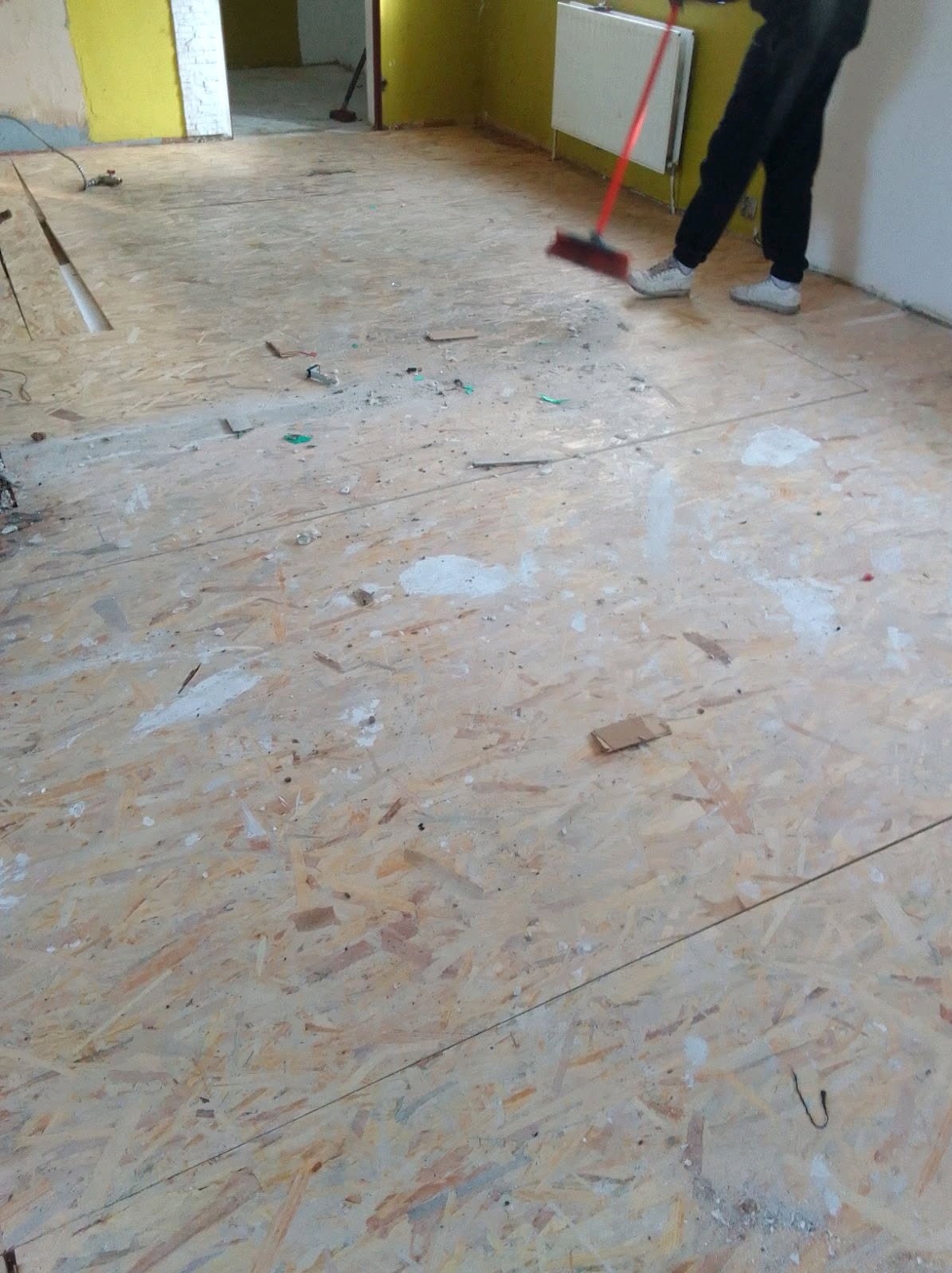Podłoga z płyt OSB w trakcie sprzątania po remoncie, częściowo posprzątana, z widocznymi resztkami gruzu i farby, osoba zamiata czerwoną zmiotką.