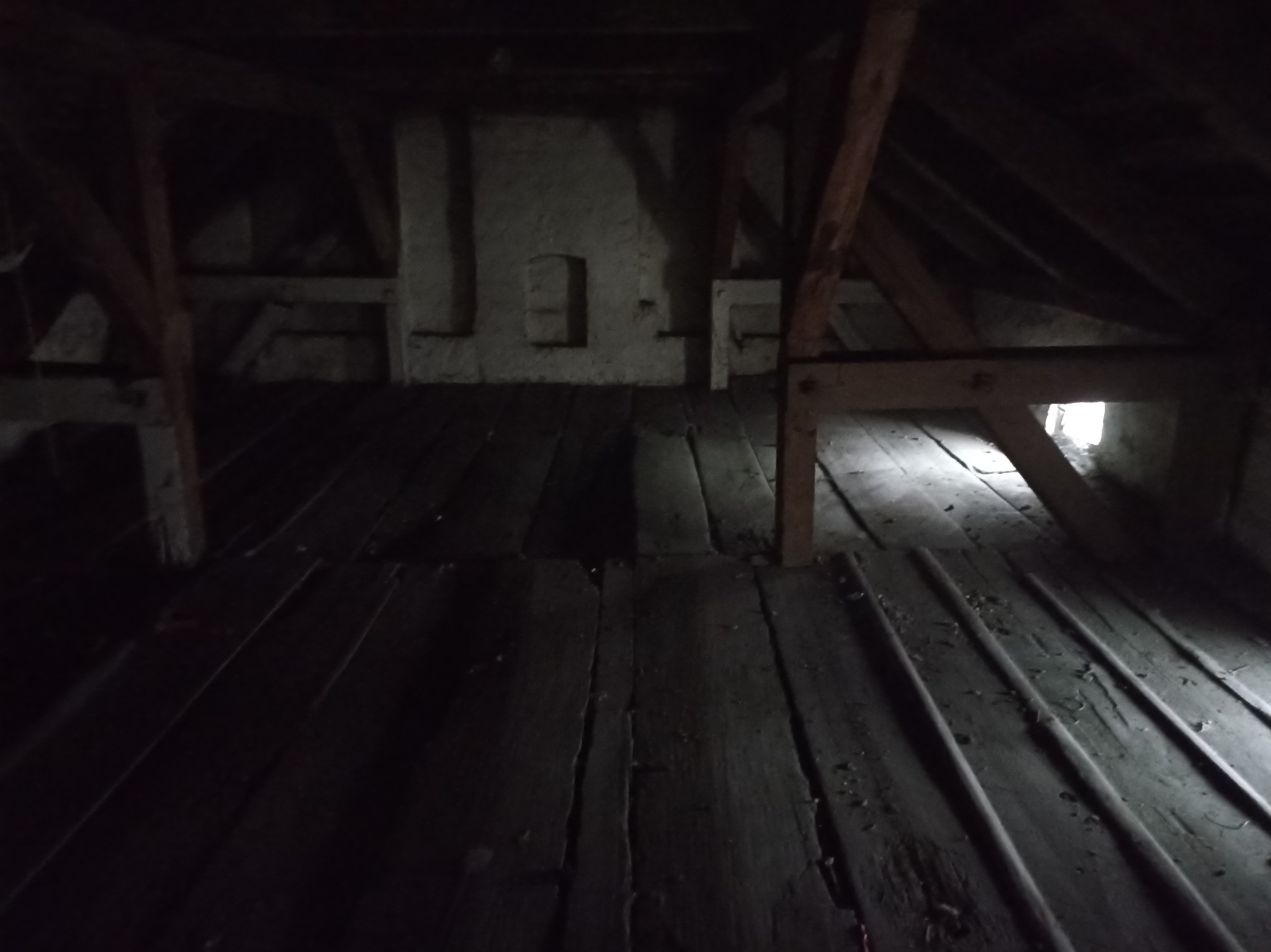 Mroczne poddasze ze starymi drewnianymi belkami i podłogą, fragment ściany z wnękami, słabe światło wpadające przez okno