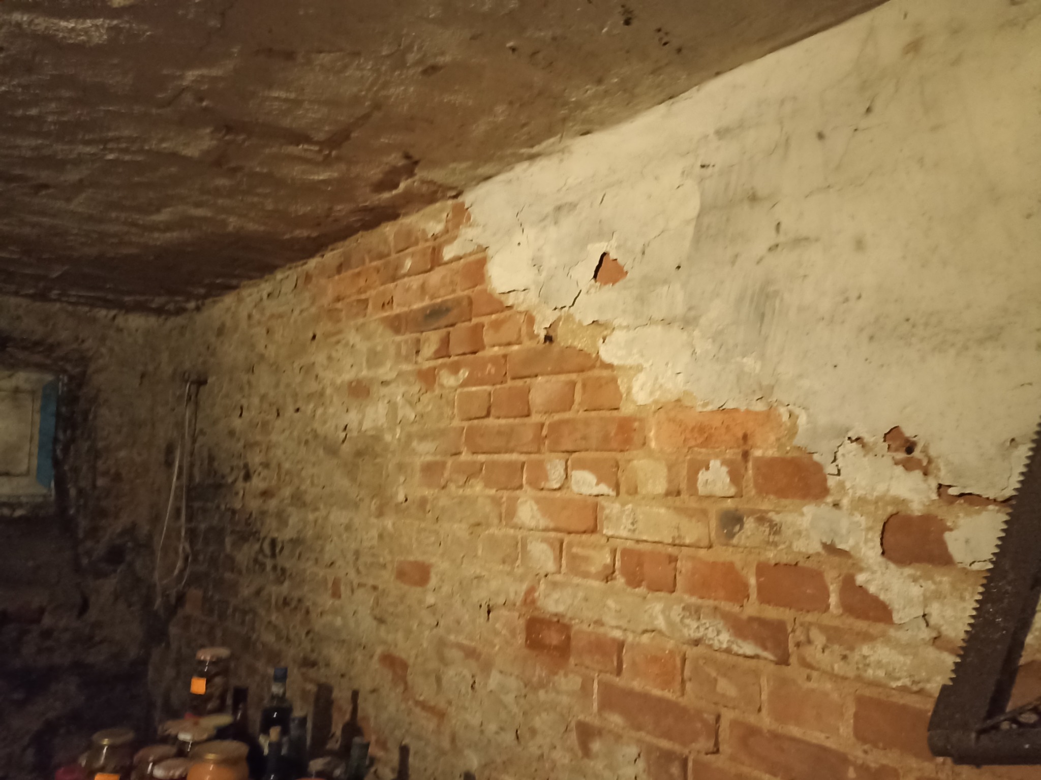 Ceglana ściana piwnicy z odpadającym tynkiem, widoczne słoiki i butelki oraz zardzewiała piła ręczna