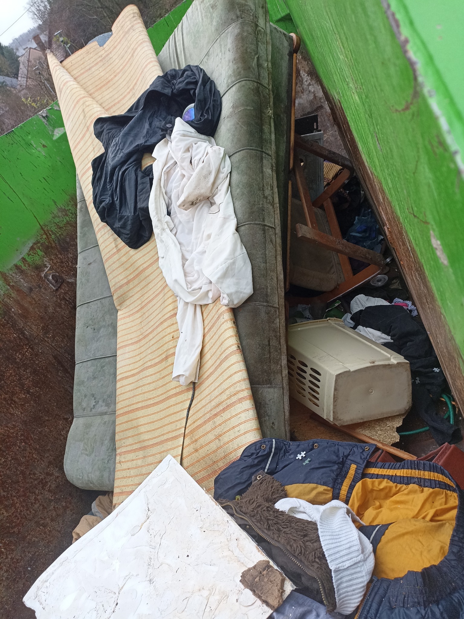 Wnętrze zielonego kontenera wypełnionego różnorodnymi odpadami: starym materacem, zwiniętym dywanem w paski, ubraniami, plastikowym koszem i fragmentami mebli.