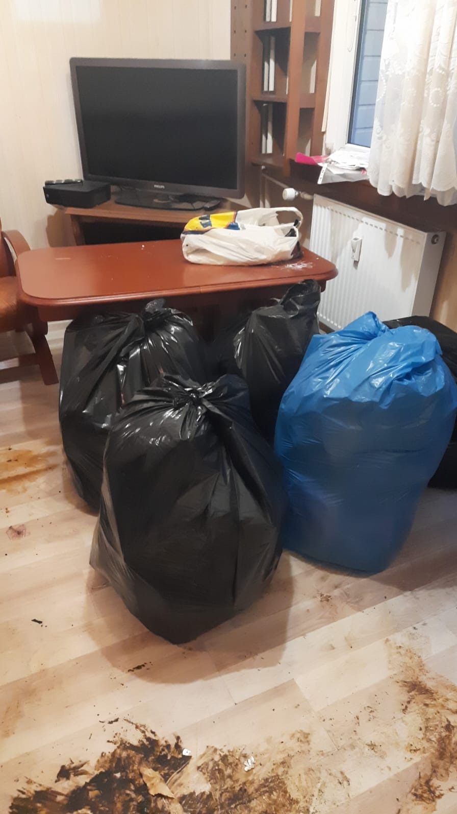 Wnętrze mieszkania z czterema workami na śmieci, trzema czarnymi i jednym niebieskim, stojącymi na zabrudzonej podłodze. W tle widoczny telewizor na komodzie i grzejnik pod oknem.