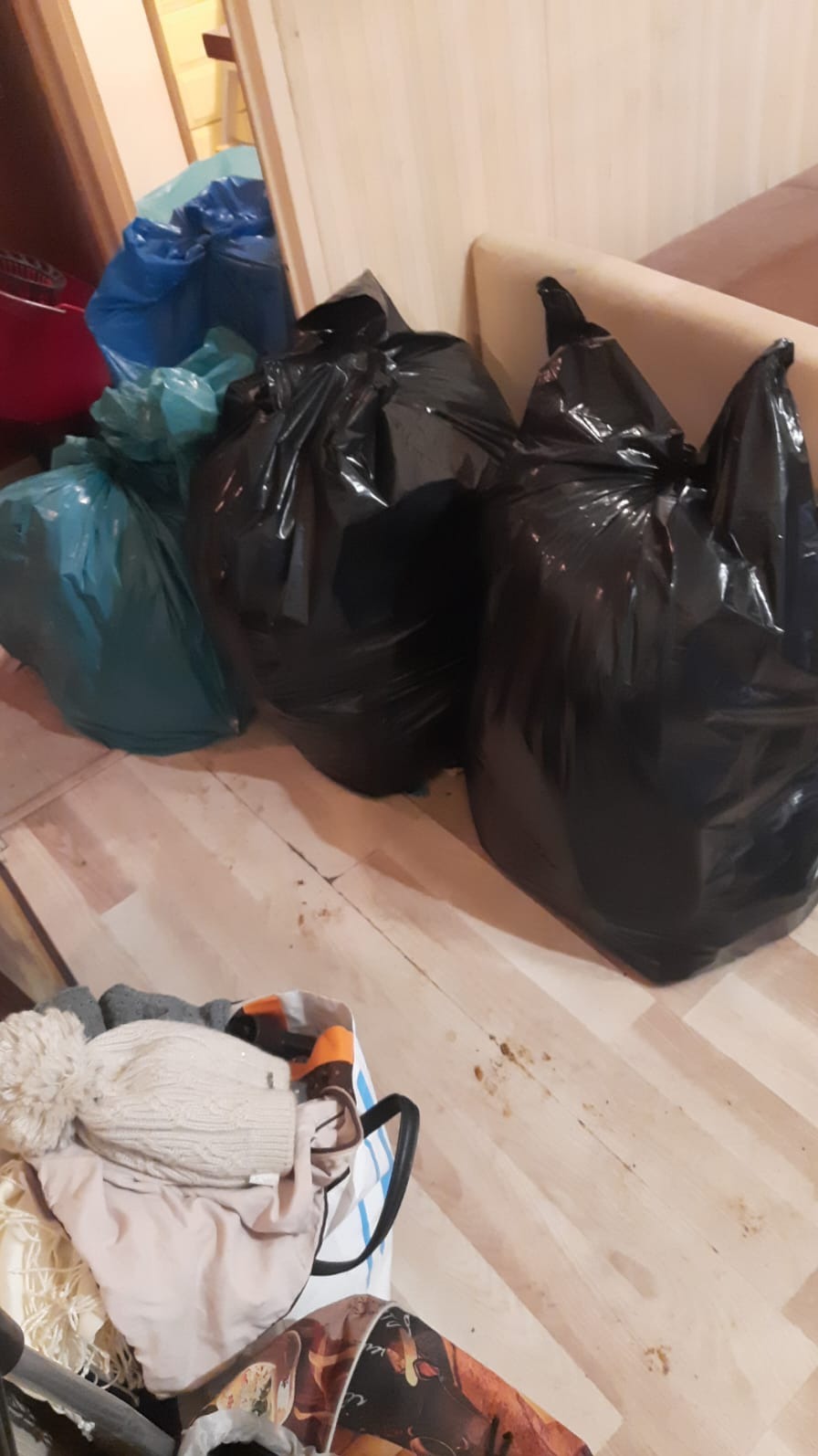 Wnętrze mieszkania z workami na śmieci w kolorze czarnym, niebieskim i zielonym, oraz torbą z ubraniami na podłodze.