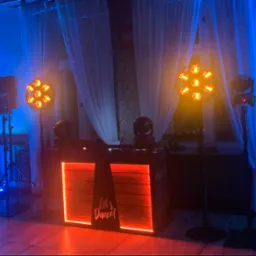 Scena DJ-a z oświetleniem scenicznym i nagłośnieniem, dekorowana w stylu retro z napisem 'Let's Dance!', w tle okno z białymi zasłonami i wzorzysta tapeta.