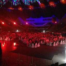 Najlepszy DJ na Imprezę Firmową, Jubileuszową. DJ Jakub - Sala widowiskowa wypełniona gośćmi siedzącymi przy okrągłych stołach, oświetlona czerwonymi reflektorami. W tle widoczna scena i konstrukcja architektoniczna.