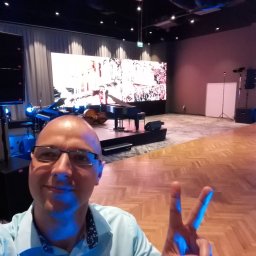 Najlepszy DJ na Imprezę Firmową, Jubileuszową. DJ Jakub - Człowiek robi selfie na tle sceny z pianinem, kontrabasem, ekranem LED i nagłośnieniem. Parkiet w jodełkę i oświetlenie sceniczne tworzą klimat imprezy.