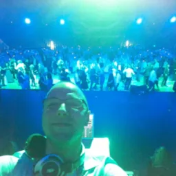 DJ robi selfie na tle bawiącego się tłumu w niebieskim oświetleniu, z widocznym sprzętem DJ-skim i słuchawkami na szyi.