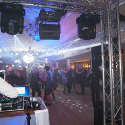 Najlepszy DJ na Imprezę Firmową, Jubileuszową. DJ Jakub - Parkiet w blasku świateł dyskotekowych, DJ za konsoletą, bawiący się ludzie w tle. Oświetlenie sceniczne zawieszone na konstrukcji kratownicowej.