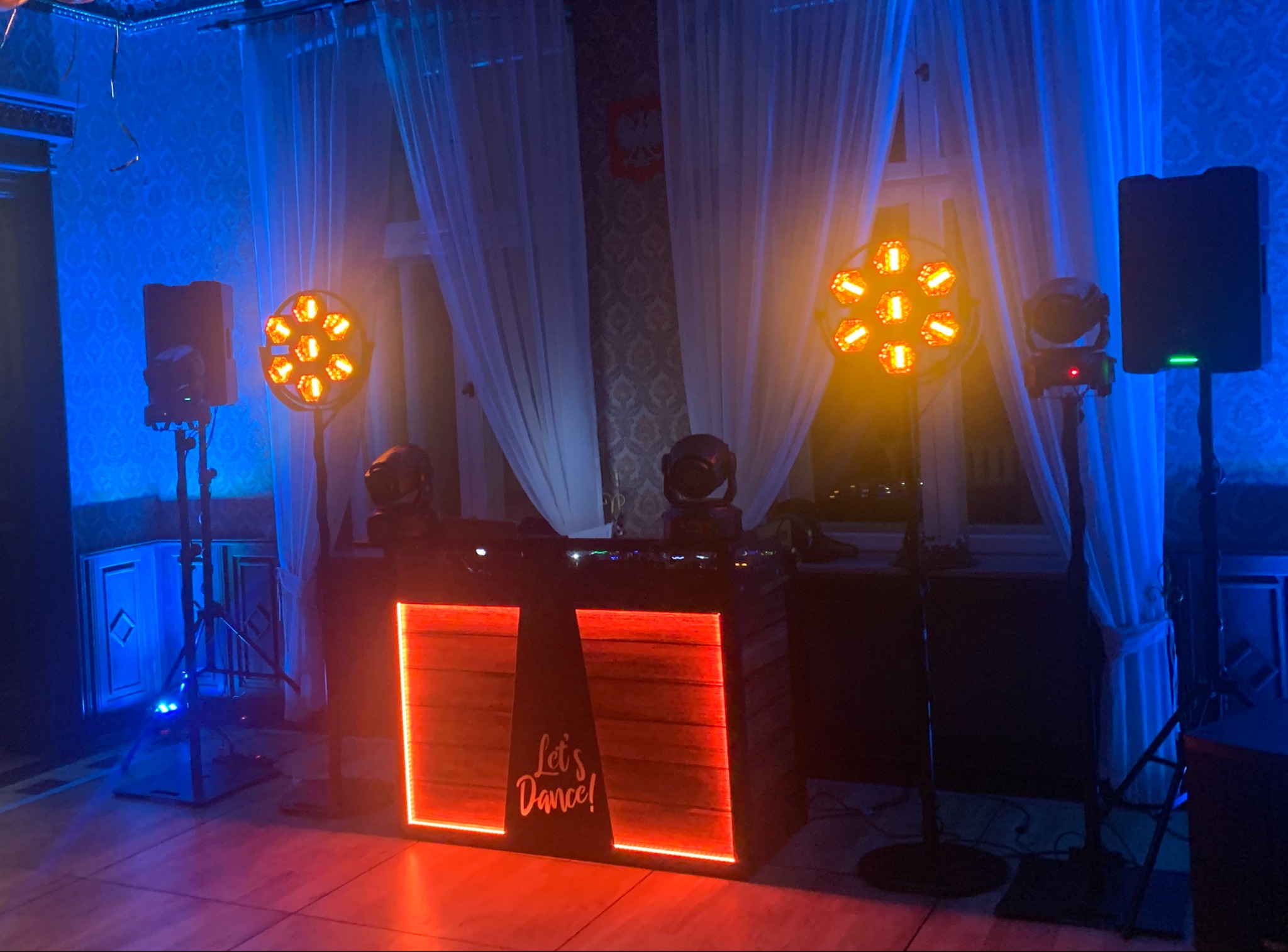 Scena DJ-a z oświetleniem scenicznym i nagłośnieniem, dekorowana w stylu retro z napisem 'Let's Dance!', w tle okno z białymi zasłonami i wzorzysta tapeta.