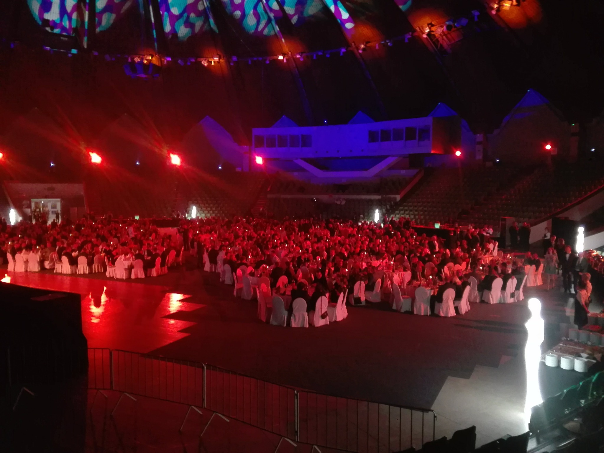 Sala widowiskowa wypełniona gośćmi siedzącymi przy okrągłych stołach, oświetlona czerwonymi reflektorami. W tle widoczna scena i konstrukcja architektoniczna.