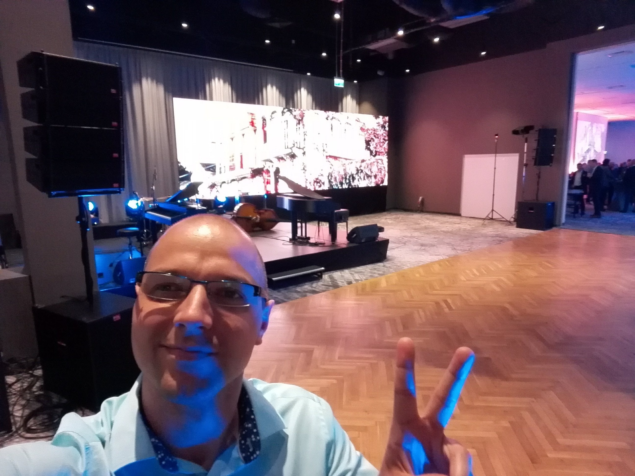 Człowiek robi selfie na tle sceny z pianinem, kontrabasem, ekranem LED i nagłośnieniem. Parkiet w jodełkę i oświetlenie sceniczne tworzą klimat imprezy.