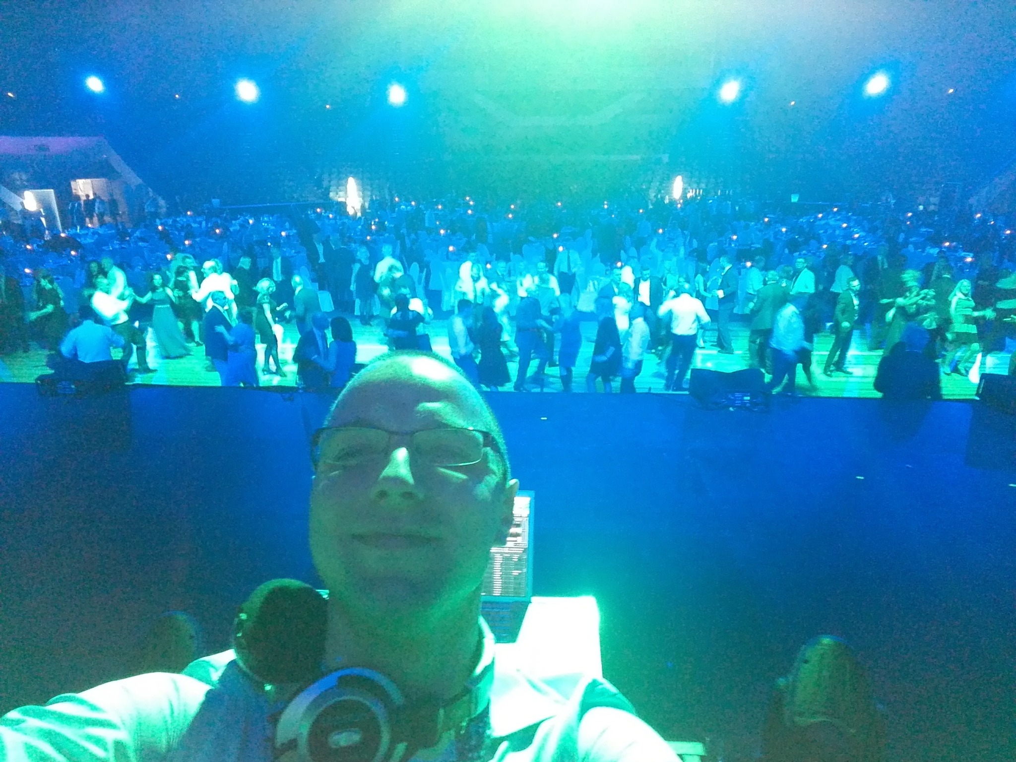 DJ robi selfie na tle bawiącego się tłumu w niebieskim oświetleniu, z widocznym sprzętem DJ-skim i słuchawkami na szyi.